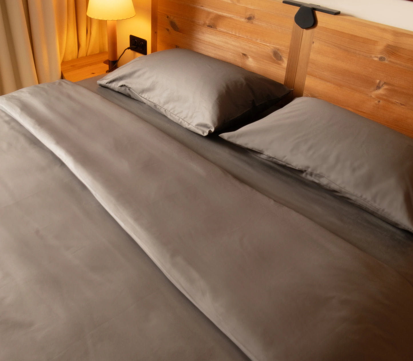 Bed with gray bedding and pillows in a room with wooden walls and a lamp.
